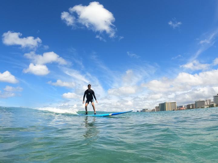 A teen standing up on a surfboard off Waikiki, with Honolulu's hotels behind