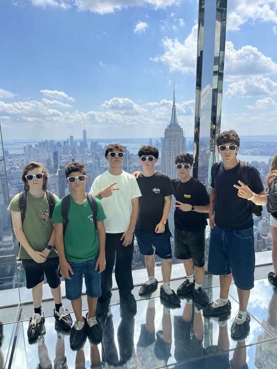 Teens standing on the glass floor of the Summit observatory, Empire State Building behind